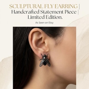 May include: A black sculptural fly earring is worn on an ear. The earring is a detailed insect design with wings and body features. Text at the top reads "SCULPTURAL FLY EARRING | Handcrafted Statement Piece | Limited Edition. As Seen on Etsy."