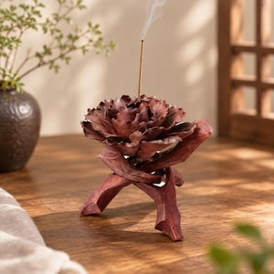 May include: A dark reddish-brown incense holder shaped like a blooming flower, resting on a tripod base made of three intersecting wooden branches. A single thin incense stick is inserted into the center, emitting a faint trail of smoke.