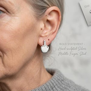 Puede incluir: Pendientes de botón 'Middle Finger' de plata esculpidos a mano. Los pendientes son una pieza audaz. El texto de la imagen dice: BOLD STATEMENT - Hand-sculpted Silver 'Middle Finger' Stud.