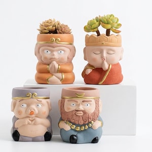 May include: Four ceramic succulent planters shaped like cartoon characters. The planters are in shades of orange, red, purple, and blue. Each planter has a different facial expression and holds a succulent plant.