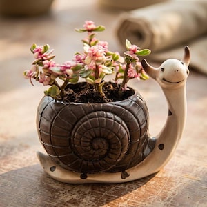 Puede incluir: Una maceta con forma de caracol de cerámica, con una concha espiral marrón oscuro y un cuerpo blanquecino con manchas negras. Contiene una pequeña suculenta con hojas rosas y verdes. Esta maceta decorativa mide aproximadamente 15 cm de largo.