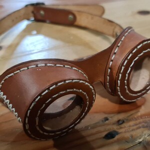 May include: Brown leather goggles with white stitching around the lenses and a matching leather strap. The goggles have a steampunk aesthetic, with a vintage look. The lenses are clear and round, and the strap has a buckle for adjustment.