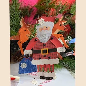 以下が含まれることがあります： 赤いチェック柄のスーツ、黒いベルト、白いひげの紙製のサンタクロースが特徴の、お祝いのクリスマスデコレーション。2頭のトナカイがサンタの両側にいます。雪の結晶のデザインの青い袋が彼の手にぶら下がっています。