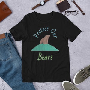 May include: Black t-shirt featuring a brown bear cub standing on a green hill. The text "Protect Our Bears" is written in blue and green lettering.