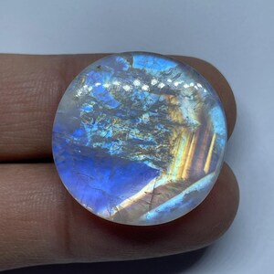 May include: A round, polished moonstone cabochon featuring a translucent surface with a prominent blue adularescence effect. The gemstone displays internal fractures and inclusions, with streaks of golden-yellow and orange light reflecting across the surface.
