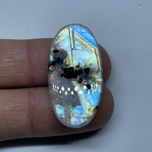 May include: An oval gemstone with a mix of colours, including blue, yellow, and black. The stone has a polished surface and appears to be a type of labradorite or moonstone. The gemstone is held in a hand, showcasing its size and shape.