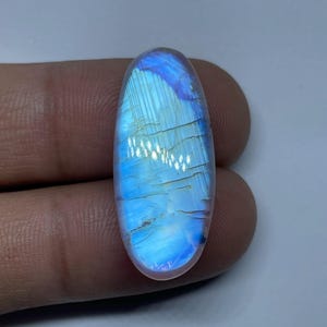 Peut inclure: Une pierre de lune ovale et polie. La pierre présente un jeu de couleurs captivant, avec des teintes bleues et blanches iridescentes. La surface présente de fines lignes et une qualité lisse et réfléchissante, idéale pour la joaillerie.