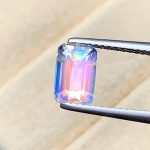 Pode incluir: Uma pedra preciosa retangular com iridescência de arco-íris, segurada por uma pinça. A gema exibe um espectro de cores, incluindo azul, rosa e amarelo, com uma superfície polida e facetada. A gema é segurada contra um fundo castanho claro.