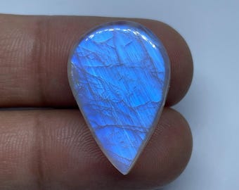Könnte beinhalten: Ein tropfenförmiger, schillernder Mondstein. Der Stein zeigt ein fesselndes Lichtspiel mit überwiegend blauer Farbe und inneren Strukturen. Die polierte Oberfläche reflektiert das Licht und erzeugt einen schimmernden Effekt. Der Edelstein wird von einer Person gehalten.