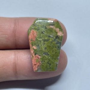 Könnte beinhalten: Ein polierter Unakit-Edelstein in rechteckiger Form. Der Stein weist ein gesprenkeltes Muster aus grünen und rosa Farbtönen auf. Der Unakit wird zwischen Fingern gehalten, wodurch seine einzigartige Farbe und Textur hervorgehoben werden. Der Edelstein ist ein natürliches Produkt.