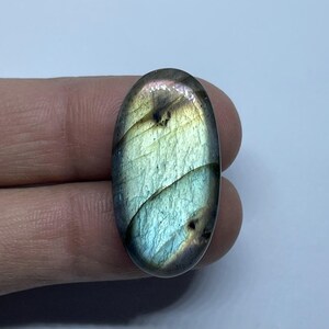 Puede incluir: Una piedra preciosa de labradorita de forma ovalada con tonos iridiscentes azules, verdes y dorados. La piedra pulida muestra un juego de colores, con una superficie lisa y reflectante. La gema se sostiene entre los dedos, mostrando su belleza natural.