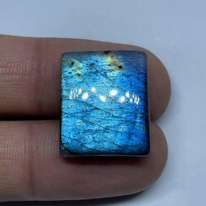 Peut inclure: Une pierre précieuse de labradorite rectangulaire avec une surface polie. La pierre présente un jeu de couleurs vibrant, avec des bleus et des verts irisés. La pierre est tenue dans une main, mettant en valeur sa beauté naturelle et ses motifs uniques.
