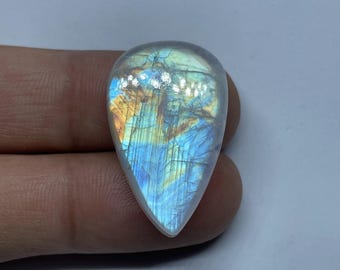 Könnte beinhalten: Ein polierter, birnenförmiger Mondstein-Edelstein. Der Stein hat eine durchscheinende, irisierende Qualität mit inneren Farben von Blau, Gold und Orange. Der Edelstein wird zwischen zwei Fingern vor einem weißen Hintergrund gehalten.