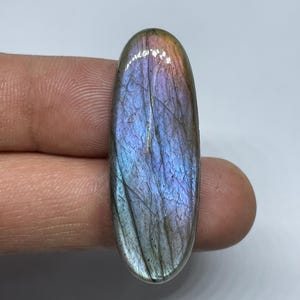 Può includere: Una pietra preziosa di labradorite di forma ovale con una superficie lucida. La pietra mostra un gioco di colori, con lampi iridescenti di blu, viola e oro. La pietra è tenuta tra due dita.