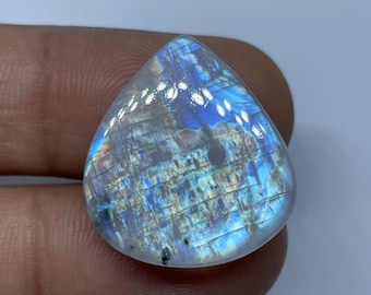 May include: A pear-shaped, iridescent moonstone gemstone. The stone displays a mix of blue, white, and gold colors with a shimmering, textured surface. The gemstone is held by a finger.