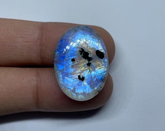 May include: An oval-shaped, iridescent moonstone gemstone. The stone displays a mix of blue, white, and black colors. The gemstone is held by a person's fingers.