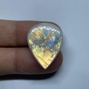 May include: A teardrop-shaped moonstone gemstone, polished to a high sheen. The stone exhibits a play of colour, with iridescent flashes of yellow, blue, and gold. The gemstone is held between fingers against a white backdrop, showcasing its natural beauty.