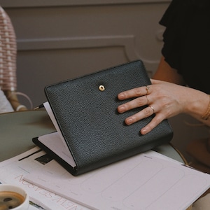 Peut inclure: Un agenda en cuir noir avec une fermeture à bouton doré, ouvert sur une page avec une mise en page de calendrier. L'agenda est sur une table avec une tasse de café et un magazine.