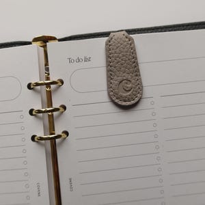 May include: A light gray leather bookmark with a debossed circular design, resting on a planner page. The page has the words "To do list" printed at the top. The planner has gold-colored rings and a dark gray cover.