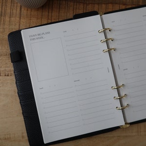 Peut inclure: Un agenda relié en cuir noir avec des anneaux dorés. L'agenda a une page blanche ouverte avec le texte "I HAVE BIG PLANS THIS WEEK..." et les jours de la semaine listés : "lund", "mardi", "mercredi", "jeudi", "vendredi", "samedi", "dimanche".