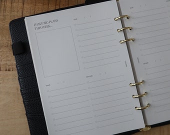 May include: A black leather bound planner with gold rings. The planner has a white page open with the text "I HAVE BIG PLANS THIS WEEK..." and the days of the week listed: "lund", "mardi", "mercredi", "jeudi", "vendredi", "samedi", "dimanche".