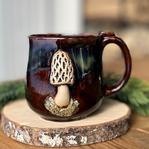 Può includere: Una tazza in ceramica marrone scuro con finitura lucida e bordo sfumato di blu. La tazza presenta una scultura tridimensionale di una spugnola di colore chiaro applicata sul davanti, con una base testurizzata che circonda il gambo.