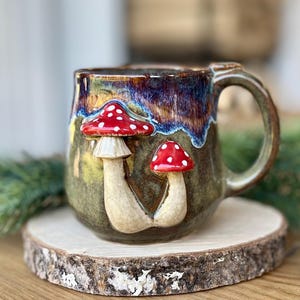 Może przedstawiać: Ceramiczny kubek z błyszczącym wykończeniem w kolorze ciemnoniebieskim i zielonym. Kubek ozdobiony jest dwoma wypukłymi, trójwymiarowymi rzeźbami grzybów z czerwonymi kapeluszami w białe kropki, umieszczonymi na zielono-niebieskim tle.