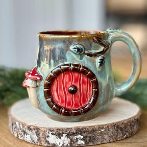 Puede incluir: Una taza de cerámica con un diseño caprichoso. La taza presenta una puerta roja, una seta y un asa de rama de árbol. La taza tiene un esmalte verde azulado y marrón y se asienta sobre una rodaja de madera.