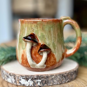 Op de afbeelding: Een keramische mok met een groene, oranje en bruine glazuur. Twee paddenstoelensculpturen zijn aan de voorkant van de mok bevestigd. De mok staat op een houten schijf.
