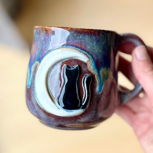 Op de afbeelding: Een keramische mok met een glanzend, meerkleurig glazuur in tinten diep paars, blauw en bruin. De mok is voorzien van een verhoogd, gebeeldhouwd ontwerp van een witte halve maan met een zwarte silhouet van een kat in het midden.
