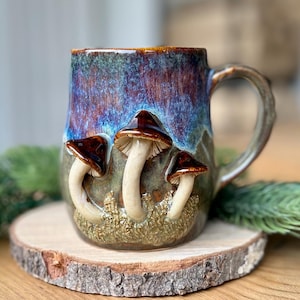 Op de afbeelding: Een keramische mok met een glanzend glazuur in tinten van diepblauw, paars en aards groen. De buitenkant is versierd met driedimensionale, gebeeldhouwde paddenstoelen met bruine hoeden en crèmekleurige stelen, die uit een textuurrijke mosbasis komen.