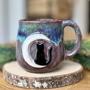 May include: A ceramic mug with a glossy finish featuring a blend of purple, blue, and green glaze. The mug displays a raised, three-dimensional white crescent moon with a black cat silhouette sitting in front of it, centred on the front surface.