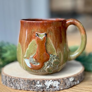 Peut inclure: Une tasse en céramique avec un motif de renard. La tasse a un glaçage marron et vert avec un renard sculpté sur le devant. La poignée et la base de la tasse sont vertes. La tasse est posée sur un dessous de verre en bois.