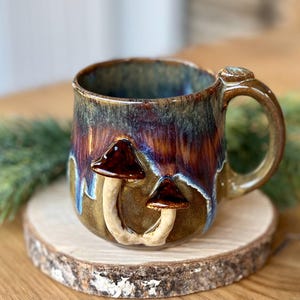 Puede incluir: Una taza de cerámica hecha a mano con un esmalte marrón, azul y dorado. Dos figuras de hongos están adheridas al exterior de la taza. La taza se encuentra sobre una pequeña rodaja de madera, con vegetación en el fondo.