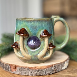 May include: A ceramic mug with a green and blue glaze, adorned with brown mushroom details and a purple stone. The mug sits on a wooden slice, with a green sprig in the background. The mug has a curved handle.