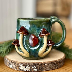 Puede incluir: Una taza de cerámica con un esmalte verde oscuro brillante que presenta setas esculpidas en tres dimensiones con sombreros marrones y tallos color crema. Una piedra pulida de color rojo parduzco está incrustada en el centro.