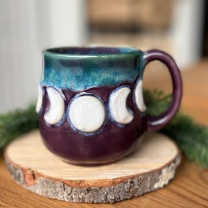 Può includere: Una tazza in ceramica fatta a mano con base viola scuro e bordo turchese. La tazza presenta un design a fasi lunari in bianco. La tazza è appoggiata su una piccola fetta di legno.