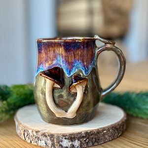 Op de afbeelding: Een keramische mok met een glanzende afwerking, voorzien van een donkerbruin en blauw druipglazuur aan de bovenzijde en een gedempte olijfgroene basis. Twee gebeeldhouwde, driedimensionale paddenstoelen met bruine hoeden en lichte stelen zijn bevestigd.