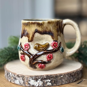 Op de afbeelding: Een keramische mok met een vogel- en bloemmotief. De mok is beige met bruine accenten en een oor. Een bruine vogel zit op een tak met rode bloemen en groene accenten. De mok staat op een houten schijf.