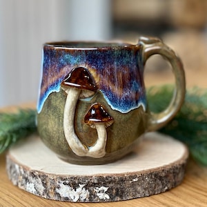 Könnte beinhalten: Eine Keramiktasse mit einem einzigartigen Pilzdesign. Die Tasse hat eine braune und weiße Pilzapplikation auf einer grün und blau glasierten Oberfläche. Der Griff hat einen passenden Erdtone. Die Tasse ist etwa 10 cm hoch.