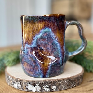 May include: A handmade ceramic mug with a unique, organic shape. The mug features a blend of colors, including brown, blue, and purple, with a glossy finish. It has a curved handle and sits on a wooden coaster. A decorative item for the kitchen.
