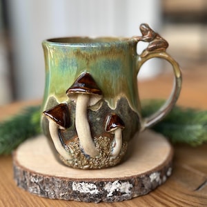 Könnte beinhalten: Eine Keramiktasse mit grün-brauner Glasur, mit dreidimensionalen Pilz- und Froschdetails. Die Tasse steht auf einer Holzscheibe und zeigt ein einzigartiges, handgefertigtes Design. Der Griff hat eine Froschfigur.
