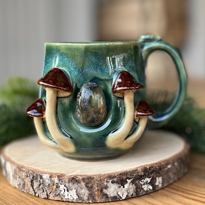 May include: A teal ceramic mug with a handle, adorned with four mushroom figures and a central stone. The mushrooms have brown caps and beige stems. The mug sits on a wooden slice, with greenery in the background.