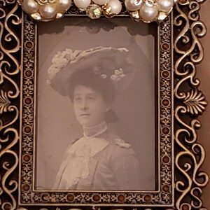 May include: Ornate, vintage-style picture frame with a sepia-toned portrait of a person wearing a hat. The frame is dark brown with gold accents, embellished with faux pearls and crystals.