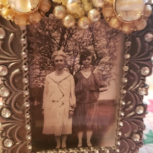 May include: Ornate, vintage-style picture frame with a sepia-toned photograph of two women. The frame is bronze-colored, embellished with clear rhinestones and decorative accents. The top of the frame is adorned with pearl-like beads and decorative embellishments.