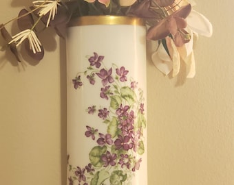 May include: A white, wall-mounted vase with a gold rim and a floral design featuring purple violets and green leaves. The vase is filled with artificial flowers in shades of pink, purple, and cream, creating a decorative display. The vase is approximately 10 inches tall.