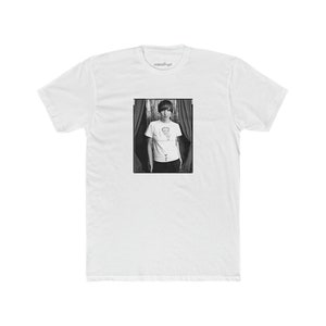 Puede incluir: Foto en blanco y negro de una persona que lleva una camiseta blanca con un gráfico de una figura de dibujos animados. La persona está de pie frente a una cortina.