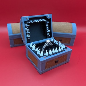 May include: A unique engagement ring box shaped like a treasure chest, with a blue frame and brown sides. The box is lined with black velvet and features white, tooth-like accents around the opening. A silver ring with a large, clear stone sits inside.