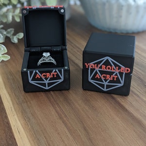 May include: A black ring box with a red and white design on the lid. The box is open and a silver ring with a large diamond is inside. The box is sitting on a wooden table next to a smaller black box with the words "You rolled a crit" printed on it.