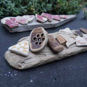 Puede incluir: Fragmentos de cerámica con motivos florales y geométricos dispuestos sobre madera flotante. La paleta de colores incluye blanco, rosa, marrón y beige. Las piezas varían en tamaño y forma, creando una exhibición rústica y artística. Medidas en centímetros.